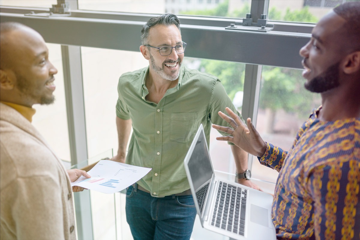 Three men having a discussion in an office, one holding a laptop and another holding a printed chart while they talk near a window.