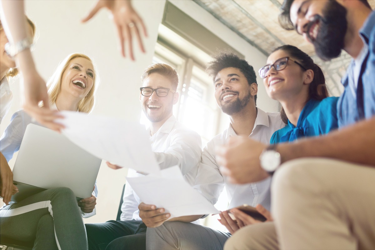 six colleagues sitting together in a bright office, smiling and laughing while reviewing papers during a collaborative team meeting.