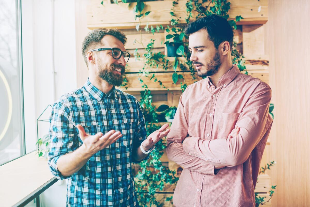 Two coworkers standing in a modern office with wooden walls and plants, engaged in conversation as one gestures while the other listens with arms crossed.