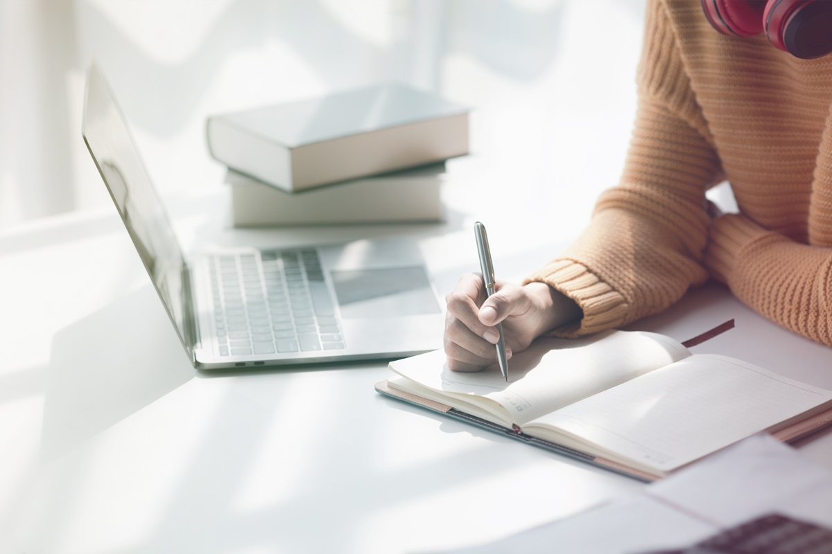 Person writing in notebook beside laptop and stacked books